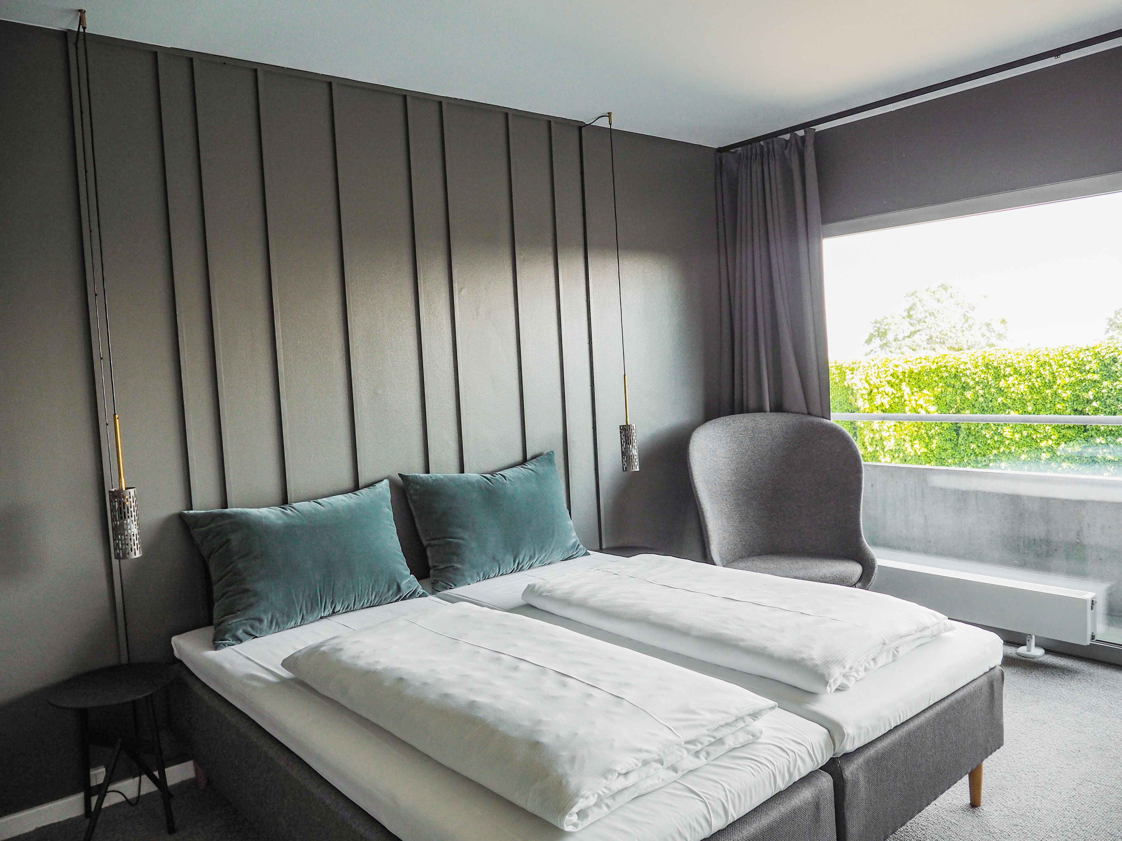 A modern hotel room with a large bed, a chair, and a view of greenery outside.
