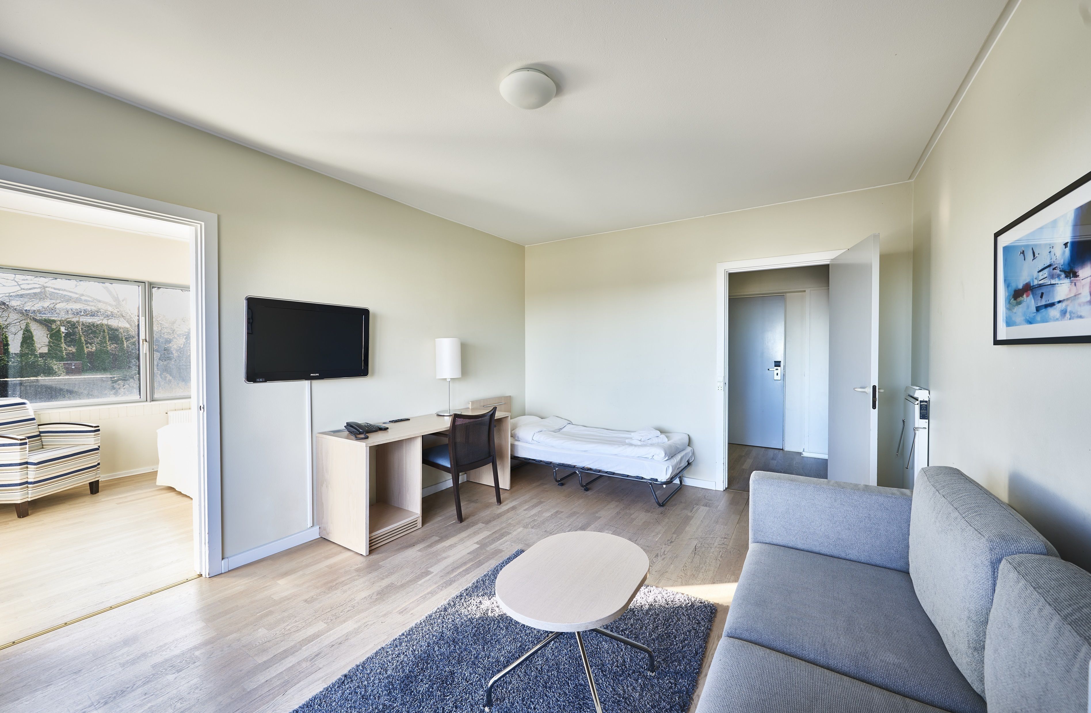 Una habitación moderna y luminosa con un ventanal grande, televisor de pantalla plana, escritorio con silla, cama, sofá y una mesita redonda pequeña.