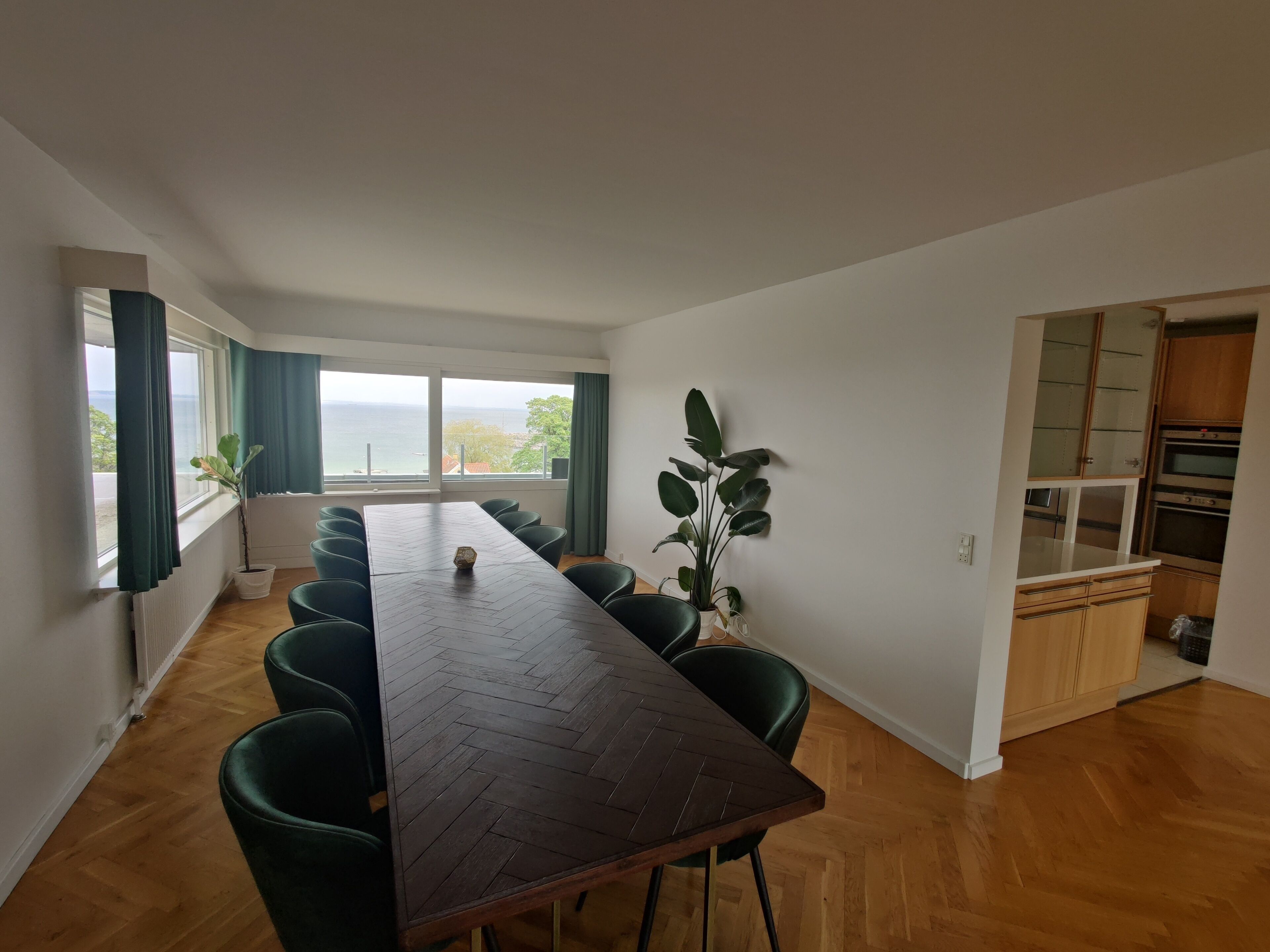 A spacious dining area with a long wooden table, green chairs, and large windows offering a view of the sea.