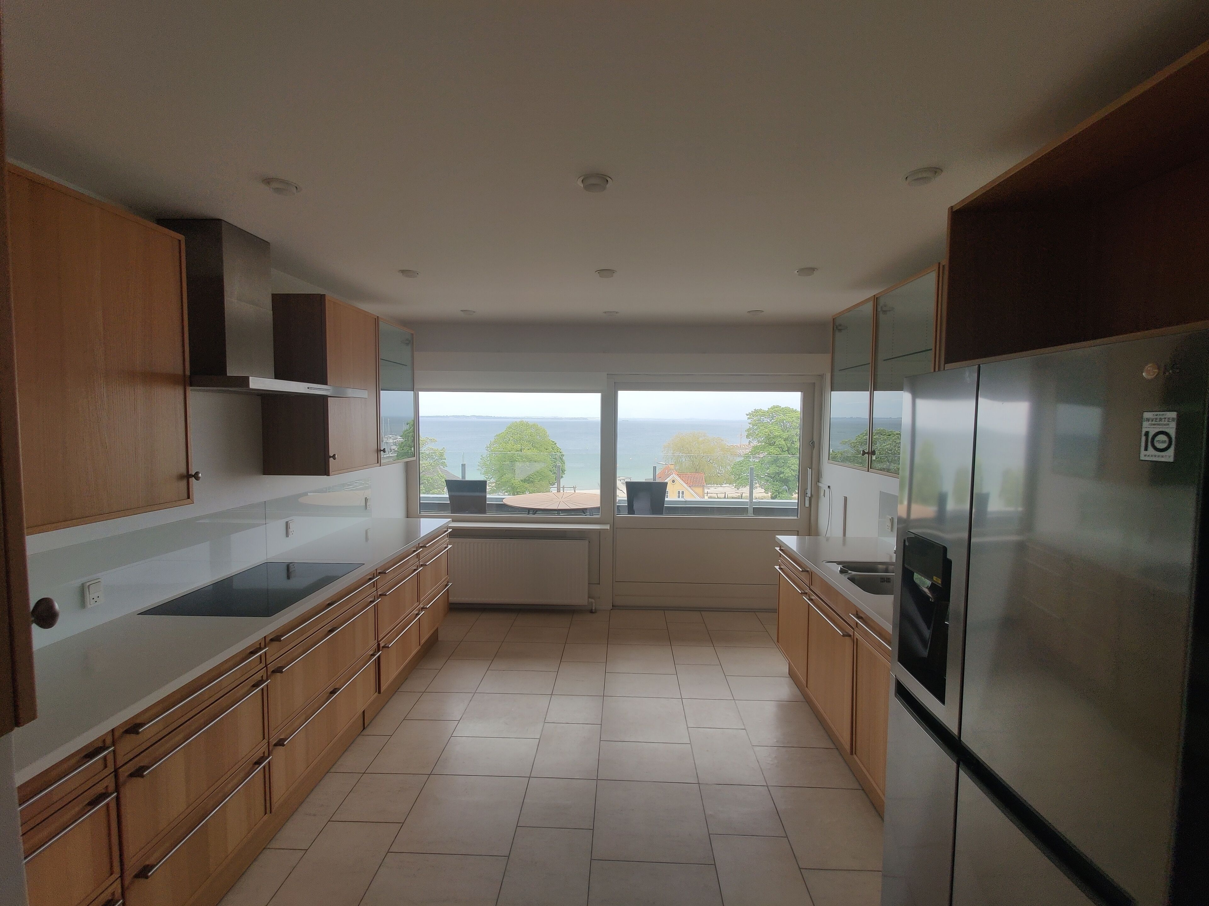 A modern kitchen with wooden cabinets, a stainless steel refrigerator, and a large window offering a view of the sea.