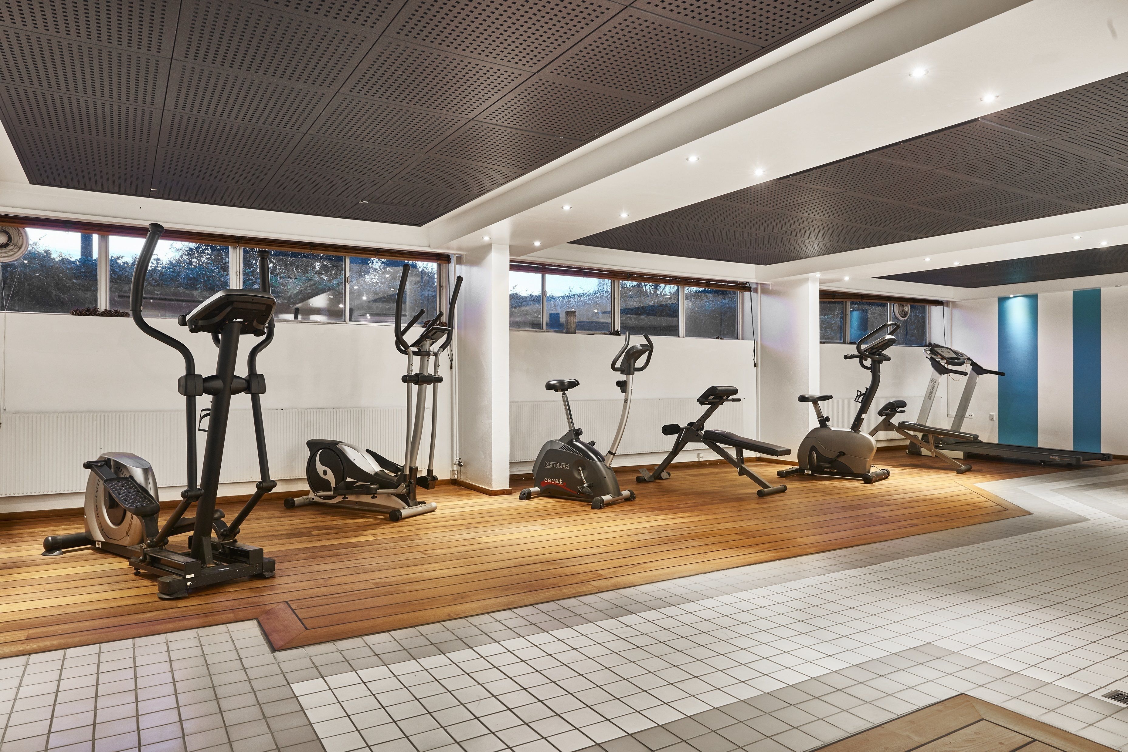 A modern gym with various exercise equipment, large windows, and a wooden floor.