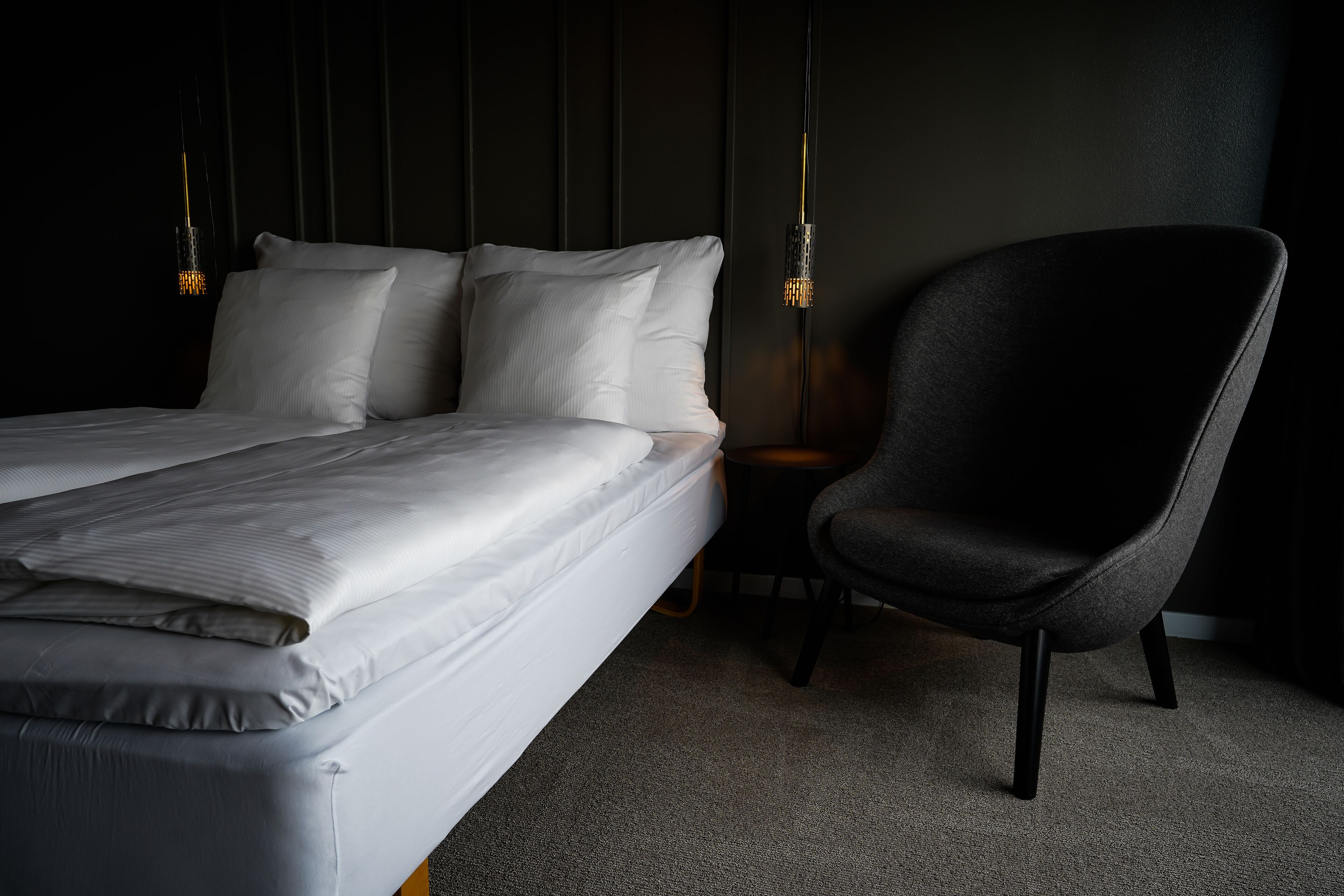 Una cama bien hecha con sábanas blancas, un sillón gris oscuro y una mesita de noche con una lámpara.
