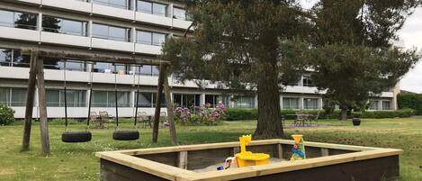 A playground with a tire swing and a sandbox in front of a modern building.
