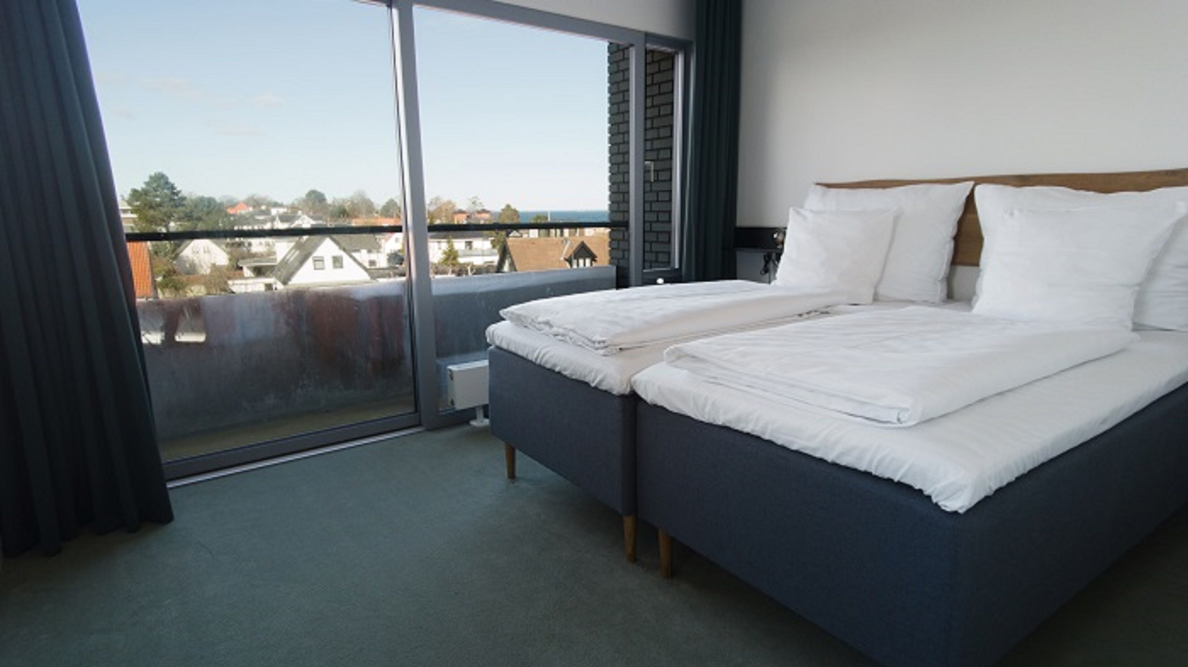 A hotel room with a large sliding glass door leading to a balcony, a bed with white bedding, and a view of buildings and trees.