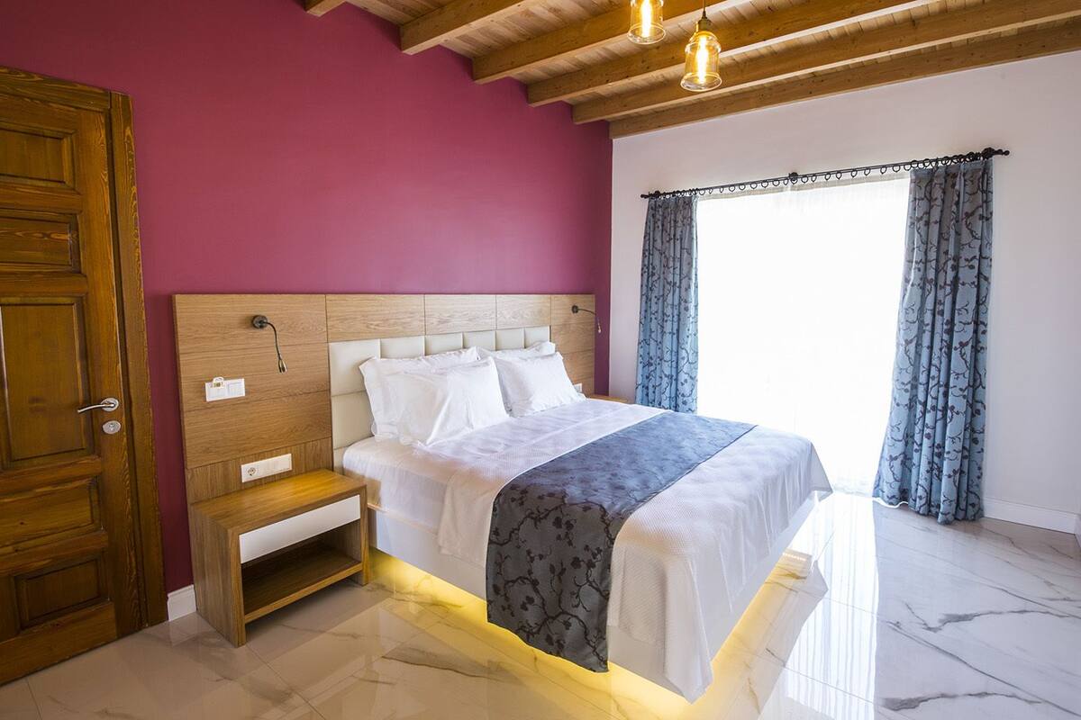 A bedroom with a wooden wardrobe, a bedside table, a bed with white bedding, a headboard, and a window with curtains.