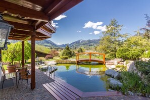 Una terraza de madera con mobiliario exterior, un pequeño estanque y un puente de madera.