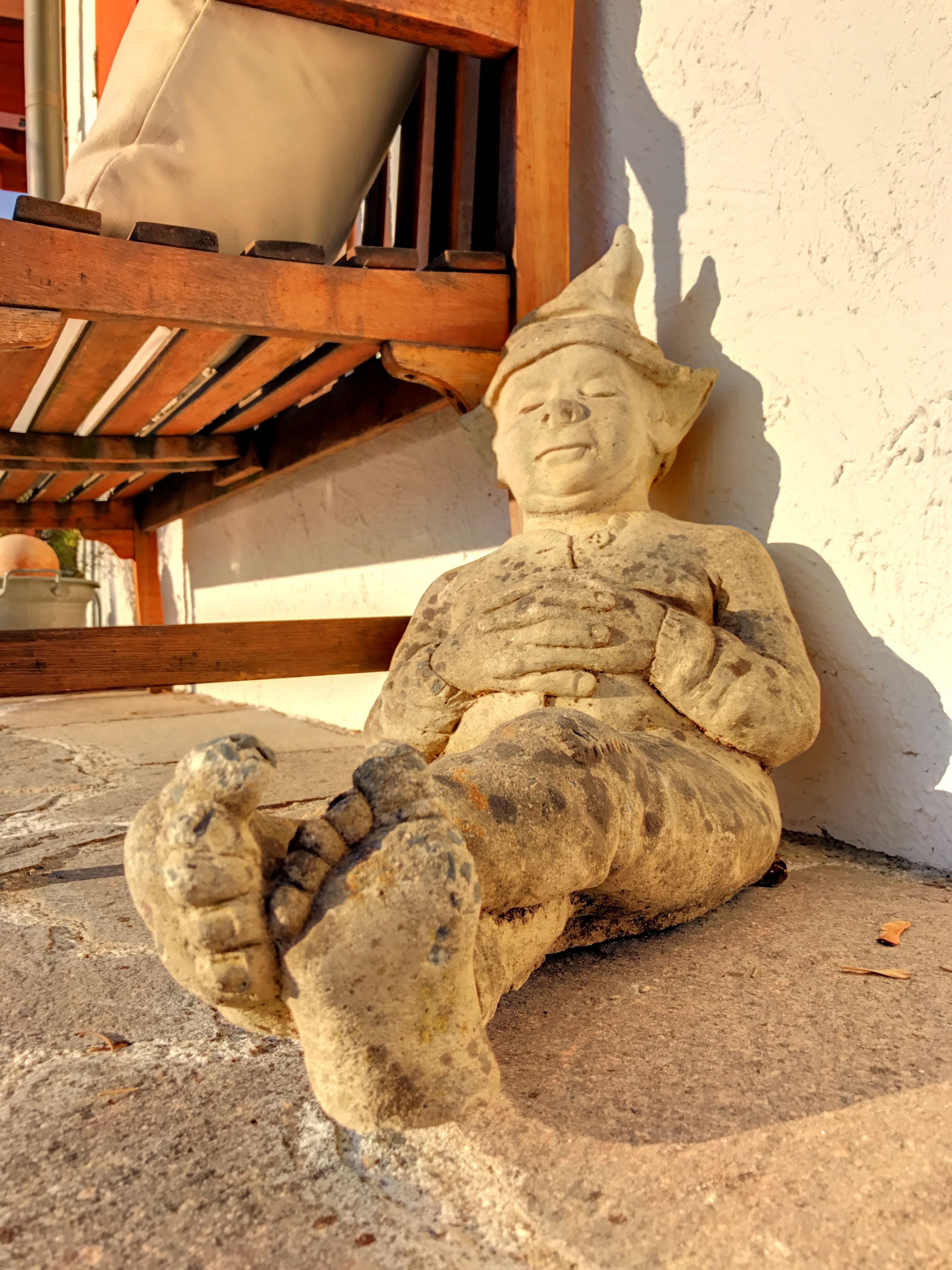 A stone statue of a seated figure with a hat, positioned against a wall with a wooden structure in the background.