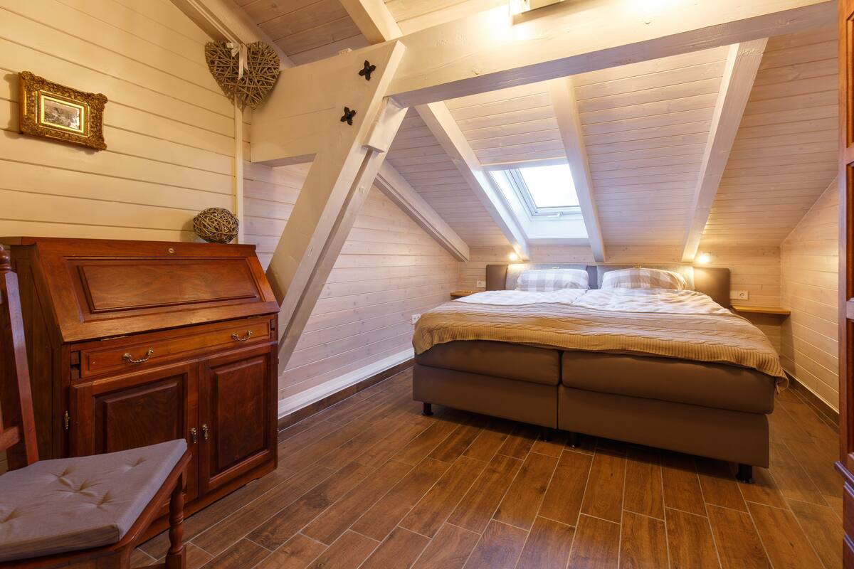 Un dormitorio con techo inclinado, suelo de madera, cama, mesita de noche, cómoda y una silla.
