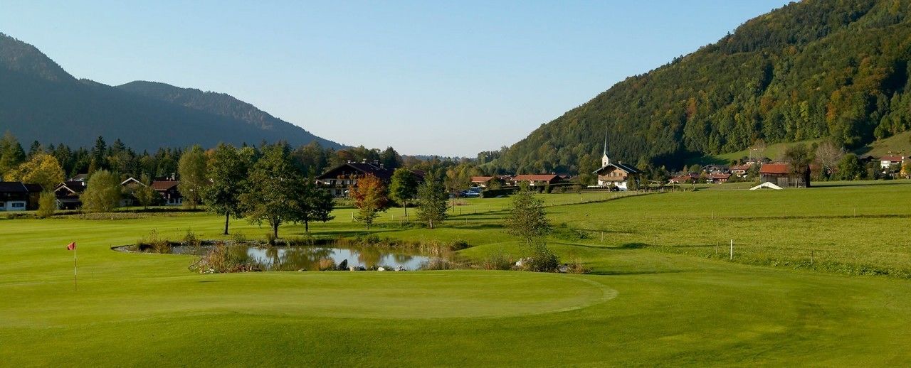 Ein Golfplatz mit Teich, Häusern und einer Kirche im Hintergrund.
