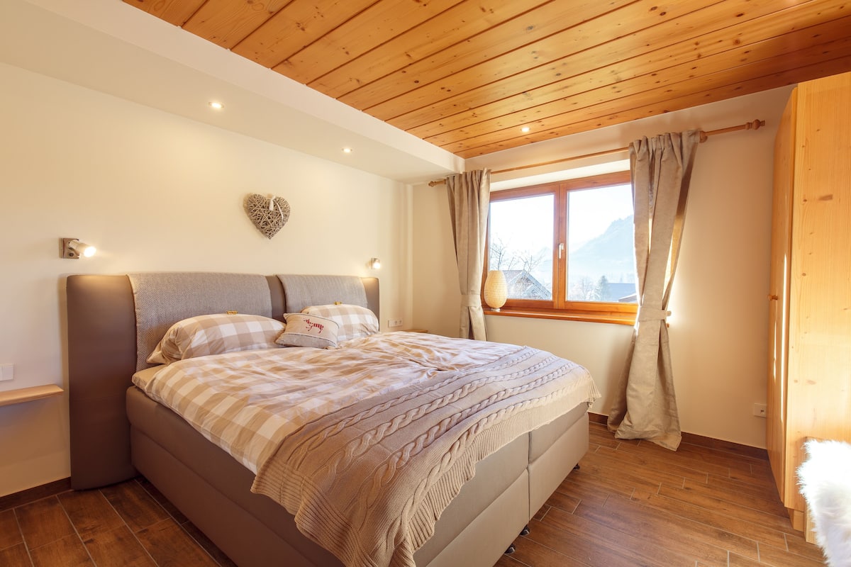 Un dormitorio con techo de madera, cama con colcha a cuadros, decoración de pared con forma de corazón y ventana con cortinas.