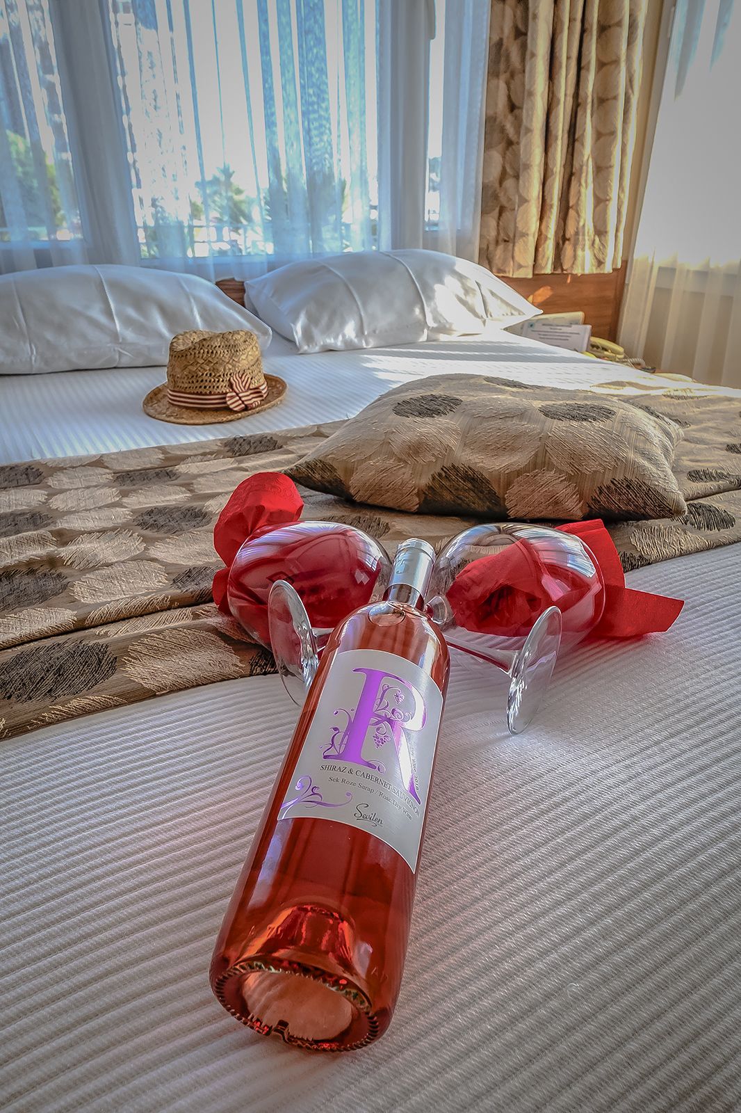A bed with a bottle of Shiraz & Cabernet Sauvignon, two red glasses, and a straw hat on a textured bedspread.
