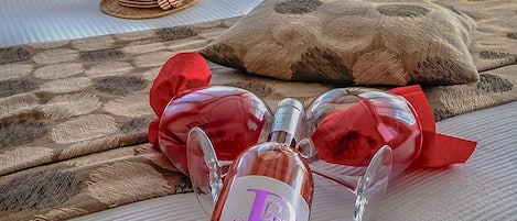 A bed with a bottle of Shiraz & Cabernet Sauvignon, two red glasses, and a straw hat on a textured bedspread.