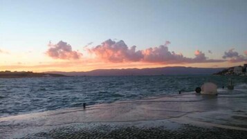A waterfront view with a sunset, calm sea, and distant mountains.