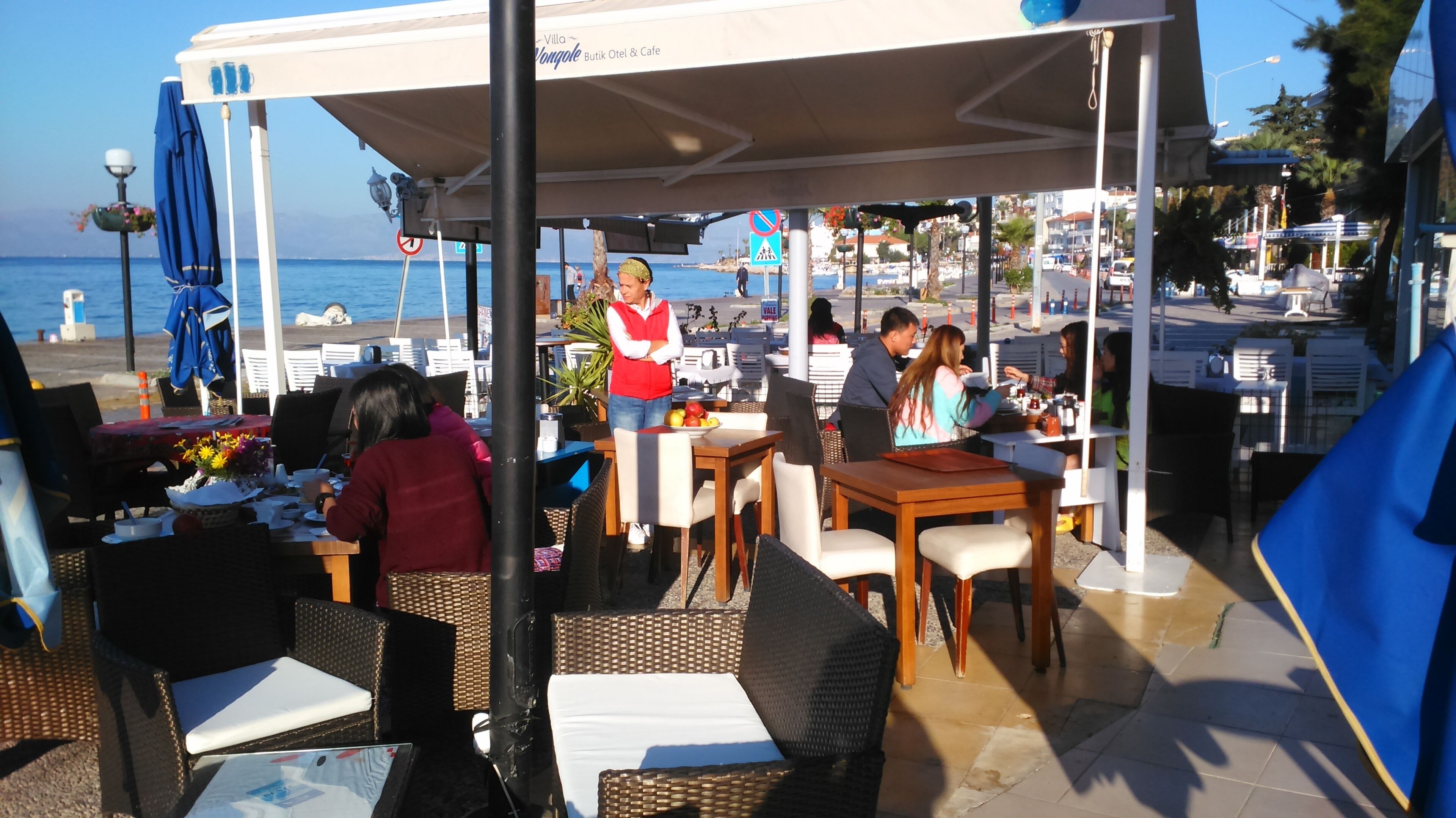 Un café al aire libre con mesas y sillas, una zona cubierta y vistas al mar.