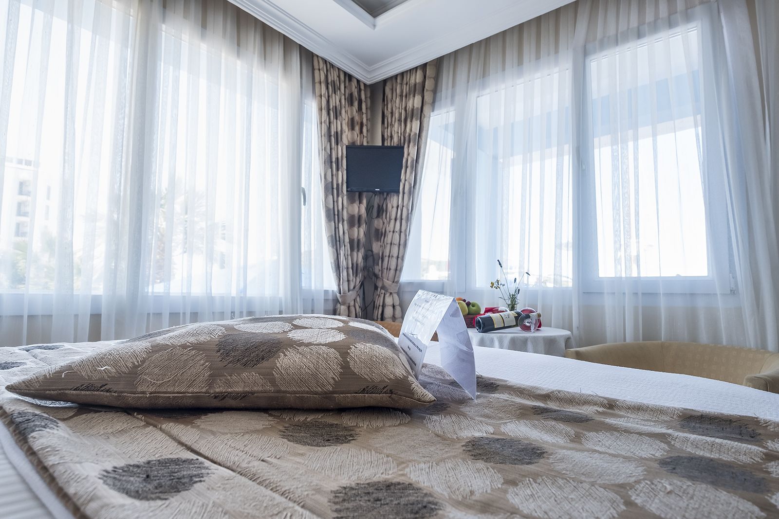 A hotel room with a bed, a small table with a vase and fruit, and sheer curtains.