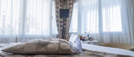 A hotel room with a bed, a small table with a vase and fruit, and sheer curtains.