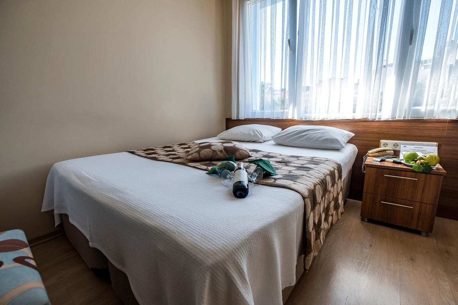 A neatly made bed with a white bedspread, a brown patterned blanket, and a bedside table with a lamp, a phone, and a fruit basket.
