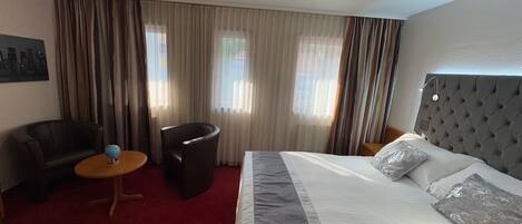 A hotel room with a bed, a chair, a small table, and curtains.