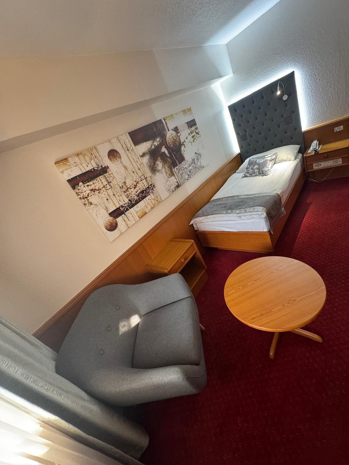A compact room with a bed, a grey sofa, a small wooden table, and a large abstract painting on the wall.