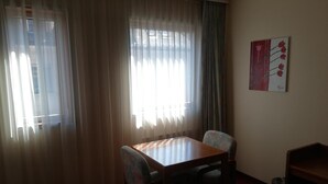A room with a wooden table and two chairs, a window with sheer curtains, and a wall-mounted painting. - Hôtel des Trois Rois (Le Locle)