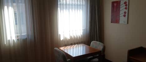 A room with a wooden table and two chairs, a window with sheer curtains, and a wall-mounted painting.