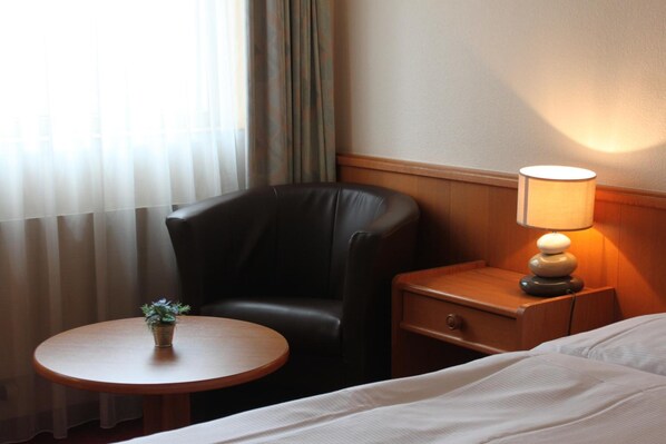 A hotel room with a bed, a small round table with a plant, a leather armchair, a wooden nightstand with a lamp, and a window with sheer curtains. - Hôtel des Trois Rois (Le Locle)