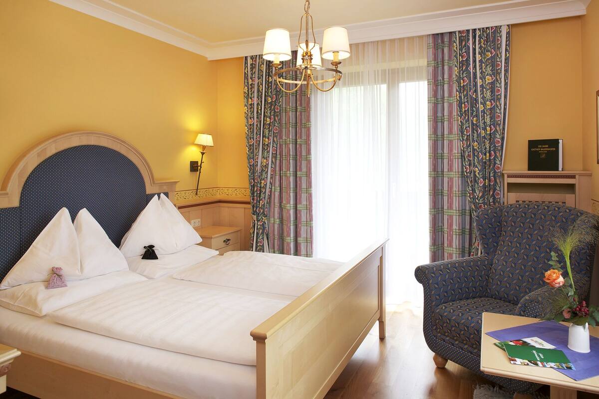 Ein Hotelzimmer mit Bett, Sessel, Nachttisch, Lampe und einem Fenster mit Vorhängen.