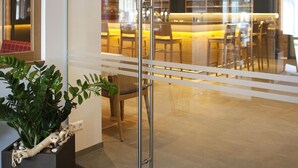 A modern interior with a glass door, a reception desk with a plant, and a view of a dining area with chairs and tables.