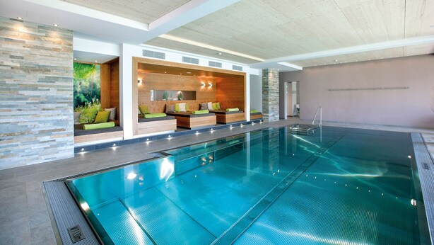 A modern indoor swimming pool with a seating area featuring wooden panels and cushions.