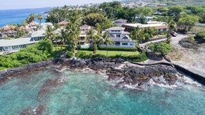 A coastal property with a clear view of the ocean, a rocky shoreline, and a well-maintained lawn with palm trees. - Ali'i Point 5 (Kailua-Kona)