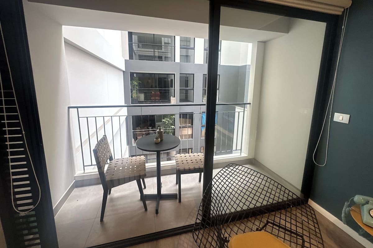 Uma varanda com mesa e cadeiras, uma pequena planta e vista para um edifício de vários andares.