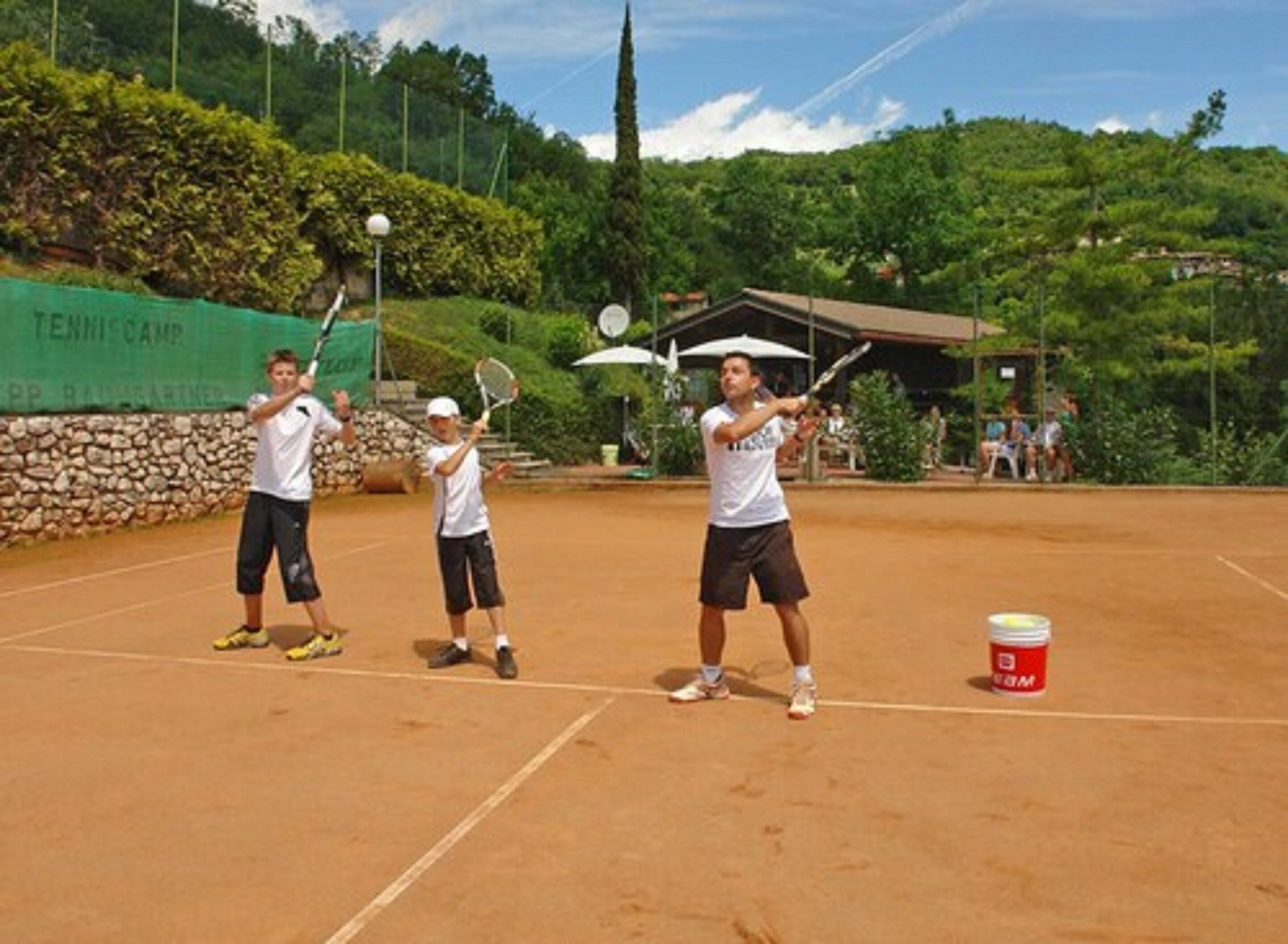 En tennisbana med tre spelare, en grön skylt och en röd behållare.