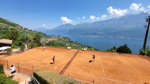 Tenniskenttä, jolta avautuu upea näkymä vuorille ja järvelle, ja jonka ympärillä on vehreää kasvillisuutta sekä asuintaloja.