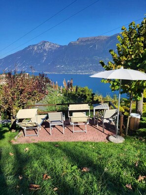 Ein Sitzbereich im Freien mit Stühlen und Tisch, einem weißen Sonnenschirm und Blick auf einen See und Berge im Hintergrund.