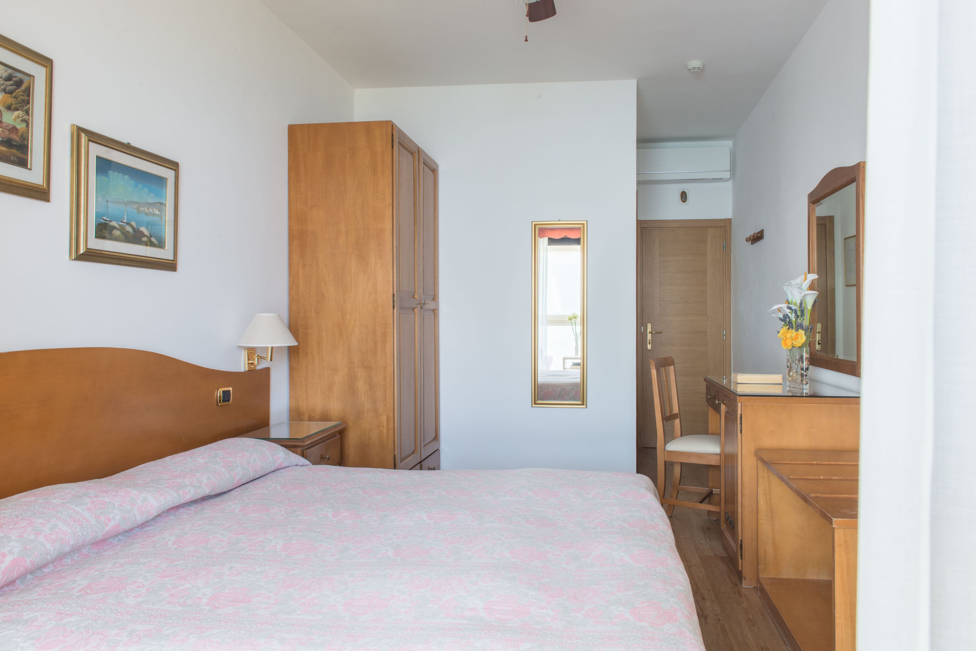 A bedroom with a bed, a wooden wardrobe, a desk with a chair, and a mirror.