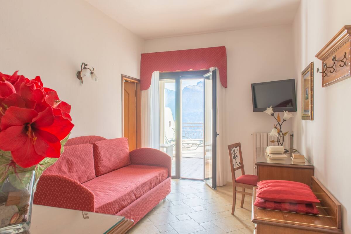 Een kamer met een rode bank, een houten stoel, een bureau met een rood kussen, een televisie aan de muur en een groot raam met uitzicht.