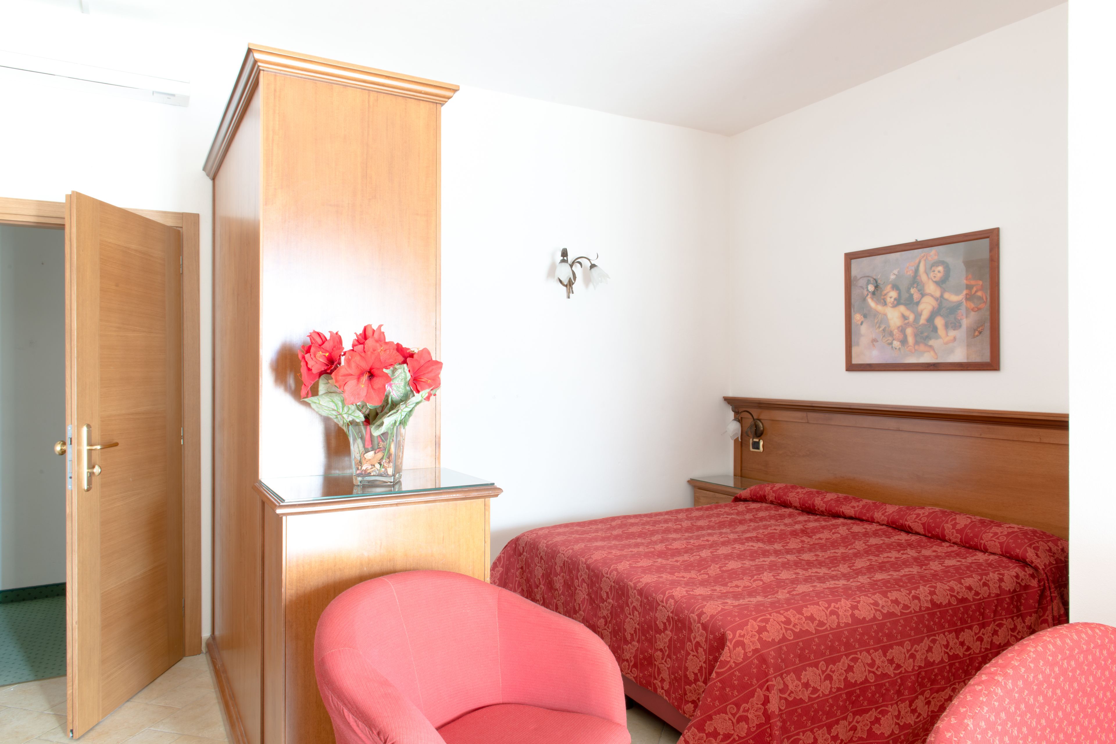 Een slaapkamer met een houten garderobe, een bed met een rode sprei, een stoel en een wandlamp.