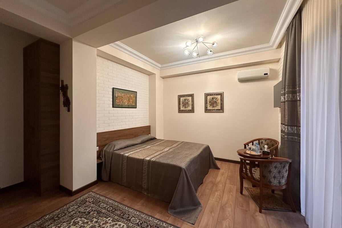 A bedroom with a bed, a nightstand, a chair, a small table, and a wall-mounted air conditioner.