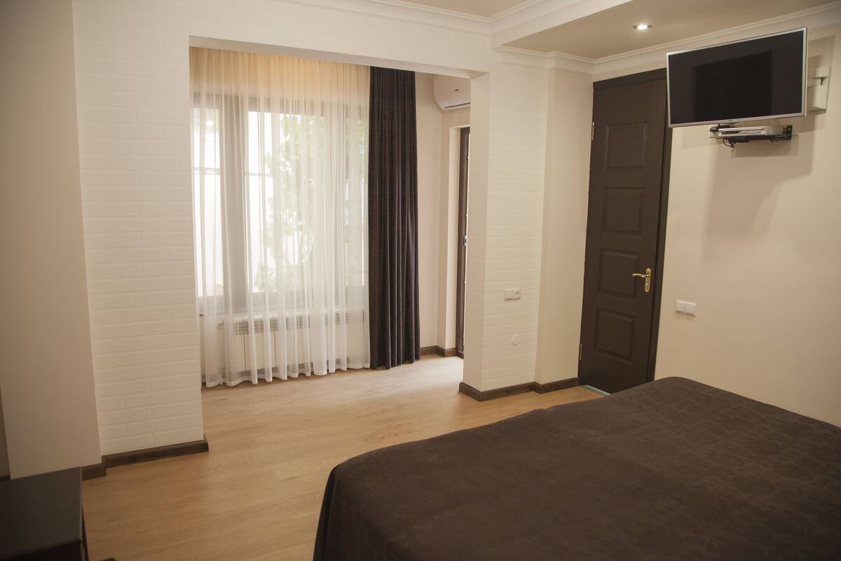 A hotel room with a bed, a TV mounted on the wall, a door, and a window with curtains.
