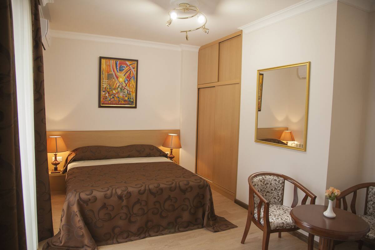 A hotel room with a bed, two bedside lamps, a chair, a table, a mirror, and a painting on the wall.