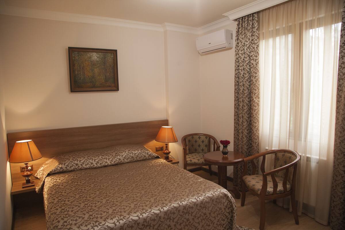 A hotel room with a bed, two lamps, a chair, a small table with a vase, and a painting on the wall.