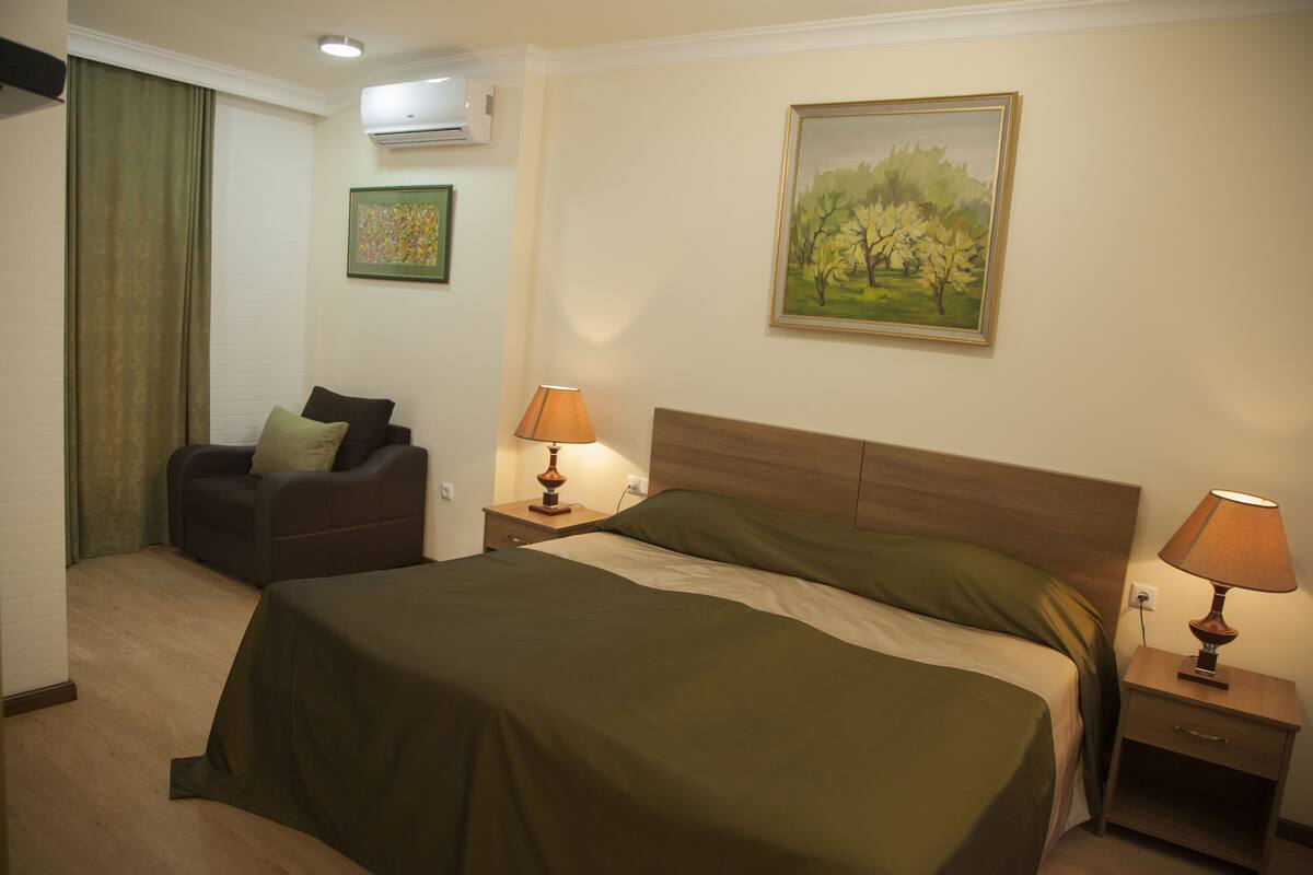 A hotel room with a bed, a nightstand with a lamp, a chair, a painting on the wall, and an air conditioning unit.