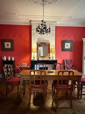 A dining room with a wooden table and chairs, a chandelier, and a red wall with a mirror and framed pictures. - Aumes Sweet Home (Aumes)