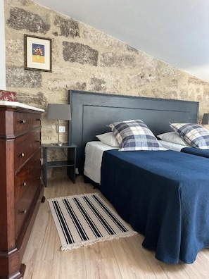 A bedroom with a stone wall, a bed with a blue headboard, a wooden dresser, and a striped rug. - Aumes Sweet Home (Aumes)