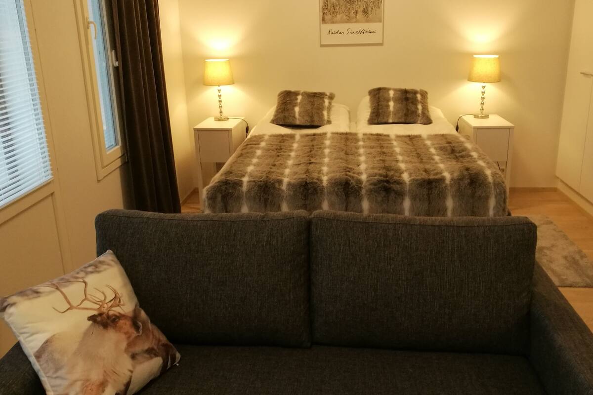 A bedroom with a bed, two bedside lamps, a window with blinds, a wall-mounted artwork, and a sofa with a decorative pillow.