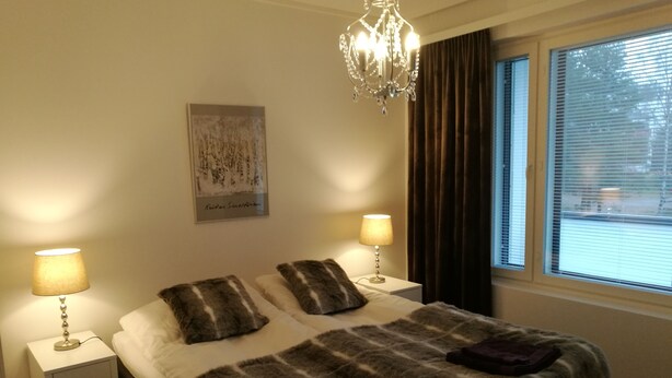 Een slaapkamer met een groot bed, twee nachtkastjes met lampen, een kroonluchter, een raam met gordijnen en een ingelijst schilderij aan de muur.