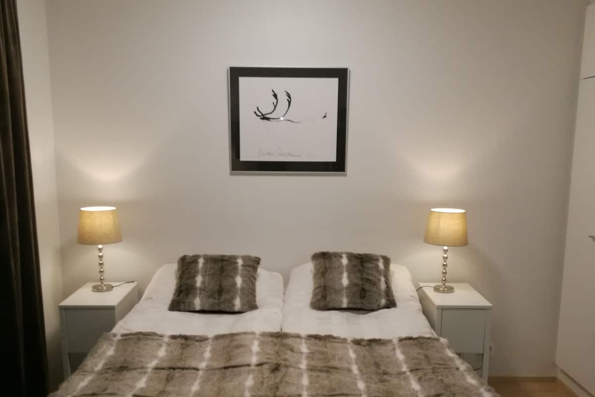 A bedroom with a bed, two bedside lamps, a framed artwork on the wall, and a window with curtains.