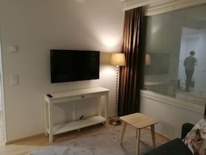 A living room with a flat-screen TV mounted on a white console, a small wooden side table, a floor lamp, and a window with curtains. - Rovavisit Apartments (Rovaniemi)