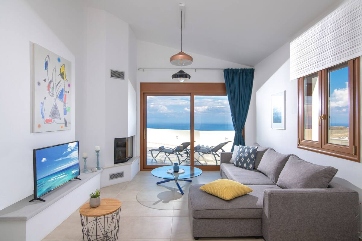 A modern living room with a large TV, a sofa, a coffee table, and a view of the beach through a sliding glass door.