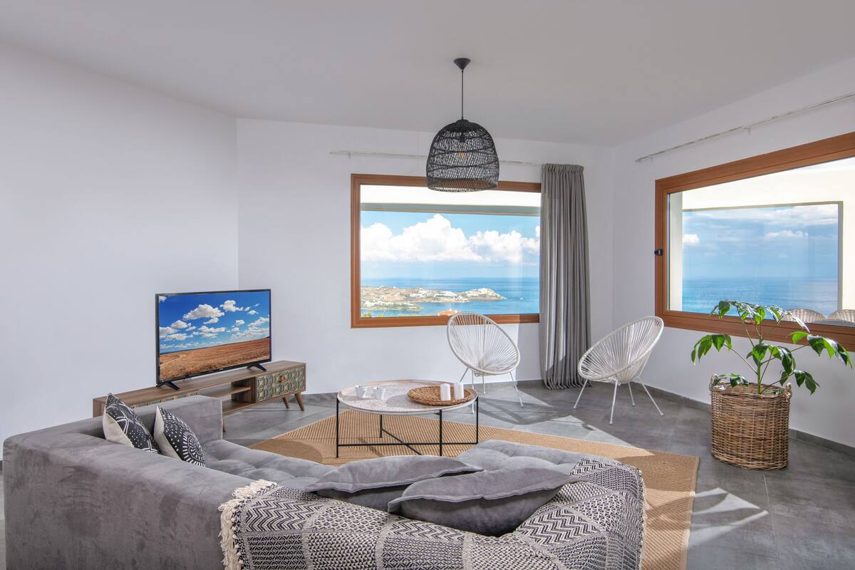 A modern living room with a large window, a flat-screen TV, a grey sofa, and a dining area.